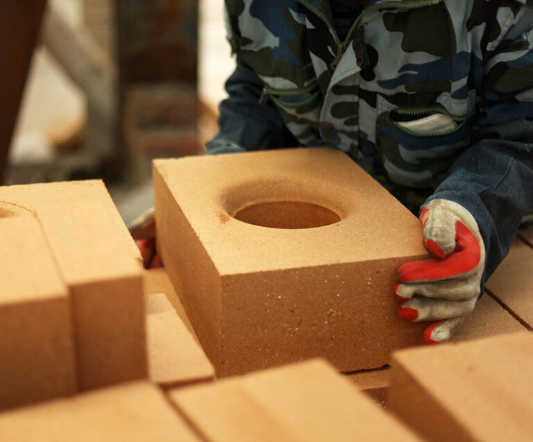 A worker wearing a camouflage jacket and red - and - white gloves is handling a tan refractory brick with a circular hole in the center. Surrounding the worker are several other similar bricks, indicating a construction or manufacturing setting.